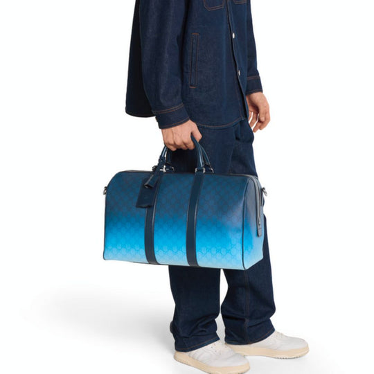 A person wearing a denim jacket and jeans carries a blue Gucci duffle bag with a gradient effect, fading from dark to light blue and featuring the GG monogram pattern. They are also wearing white sneakers.