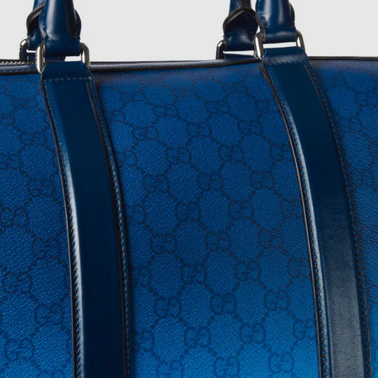 A close-up of a blue Gucci duffle bag featuring a gradient color effect, the GG monogram pattern, and dark blue leather straps.