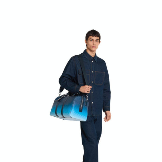 A person wearing a denim shirt and matching jeans stands with a blue Gucci duffle bag slung over their shoulder. The bag has a gradient effect, transitioning from dark to light blue, and features the repeating GG monogram pattern.