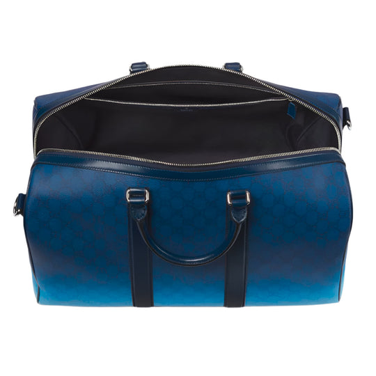 An open blue Gucci duffle bag, seen from above, displaying its dark interior, gradient blue color, GG monogram, and leather handles.