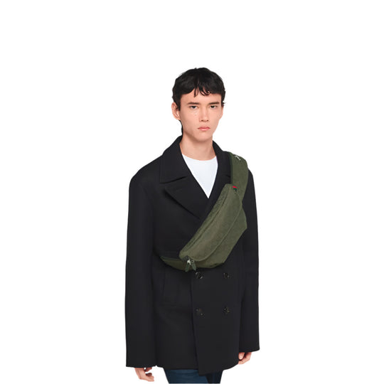 A person with short, dark hair, wearing a dark double-breasted coat over a white t-shirt, stands facing forward. A forest green Gucci GG Canvas Large Belt Bag with the signature red and green stripe detail is worn across their chest. The background is solid white.