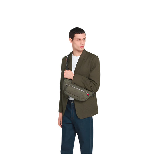 A fair-skinned man with short dark hair, wearing a dark olive green blazer over a white t-shirt and blue jeans, holds the GUCCI GG Canvas Large Slingbag across his chest. He is looking towards his right, slightly away from the camera. The slingbag is made of forest green GG canvas and features a red and green Web detail on the right side.