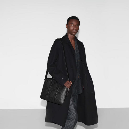 A male model with short dark hair, wearing a long black coat, a patterned dark grey vest, and matching patterned grey trousers, stands against a plain white background. He is looking directly at the viewer and holding a black Gucci GG Jumbo Medium Briefcase with his right hand, which is resting on his right thigh. The briefcase features a prominent embossed GG pattern and has a shoulder strap. The lighting is bright and even.