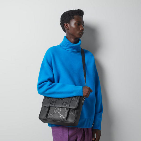 A male model in a blue turtleneck and purple pants holds a black Gucci GG Jumbo Medium Messenger Bag with embossed GG logos.