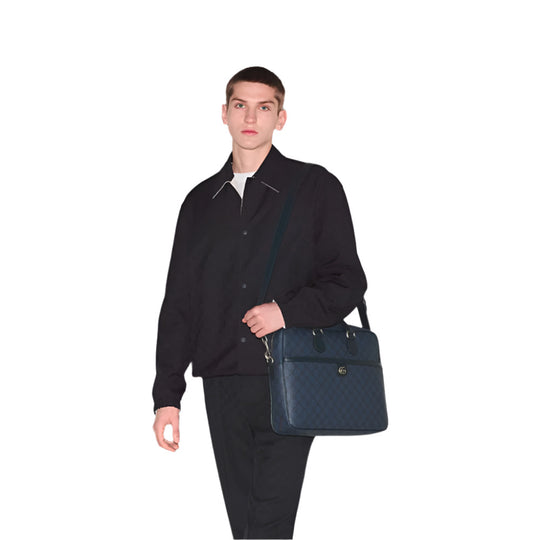 A man in a dark blue jacket and trousers holds a dark blue Gucci Ophidia Medium GG briefcase against a white background.