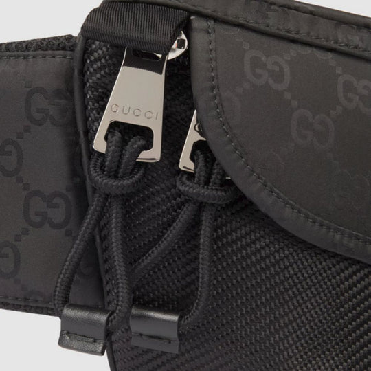 A close-up shot of a black Gucci belt bag, showcasing the tonal GG monogram pattern embossed across the nylon fabric. The silver-toned zipper pull is prominent in the center, engraved with the "GUCCI" logo. Black drawstring pulls are visible beneath the zipper, adding a utilitarian detail. The textured fabric of the bag and the smooth finish of the zipper are clearly visible, highlighting the brand's attention to detail.