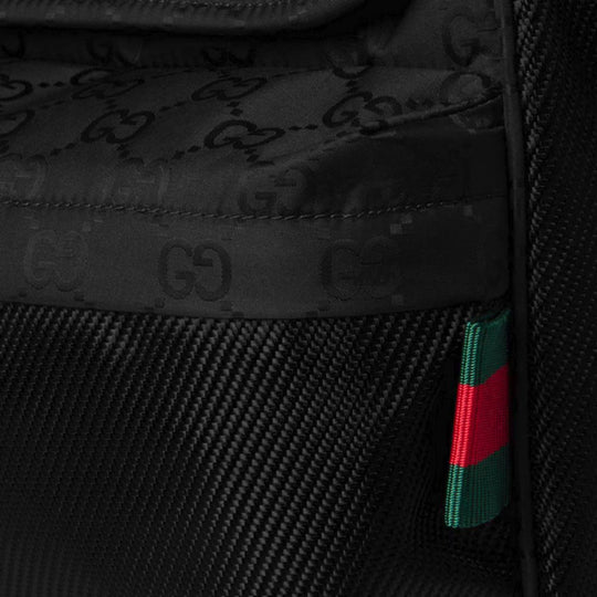 A close-up of the black GUCCI Nexus Medium Crossbody Bag, showing the tonal GG monogram on the fabric above a textured panel, and a small red and green Web stripe tag on the side.