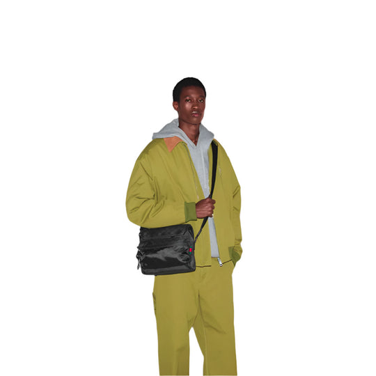 A person with short dark hair is shown holding a black GUCCI Nexus Medium Crossbody Bag with a red and green stripe detail. They are wearing a light green jacket over a grey hoodie, standing against a white background.