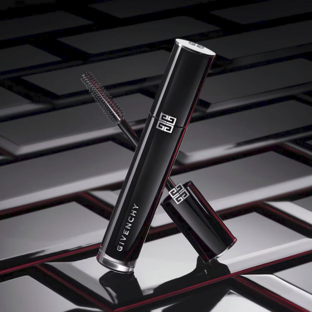 A sleek, glossy black tube of Givenchy L'Interdit Mascara Couture Volume is angled diagonally against a dark, textured background with subtle red accents. The silver cap is slightly open, revealing the mascara wand with its brush. The Givenchy logo is prominently displayed in silver on the tube. The overall impression is luxurious and sophisticated, highlighting the product's design.