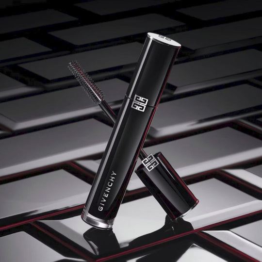 A sleek, glossy black tube of Givenchy L'Interdit Mascara Couture Volume is angled diagonally against a dark, textured background with subtle red accents. The silver cap is slightly open, revealing the mascara wand with its brush. The Givenchy logo is prominently displayed in silver on the tube. The overall impression is luxurious and sophisticated, highlighting the product's design.