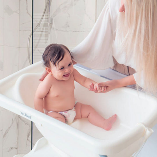 Baby sitting on Jikel Ripple Bathing Table seat with a woman holding their hand in a bathroom setting.