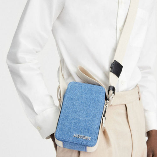 A denim blue Jacquemus Vertical Cuerda cross-body bag with a cream strap and trim, worn by a person in a white long-sleeved shirt and light-colored trousers.