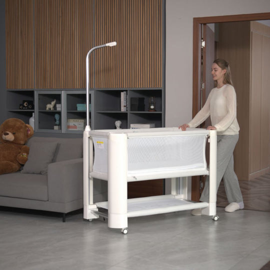A woman pushes the Jikel Braha Intelligent 7-in-1 Light Beige Baby Crib, which has a sleek white frame, mesh sides, and an attached lamp.