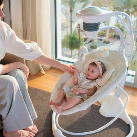 Baby in a white baby swing with a woman sitting beside it, in a room with large windows.