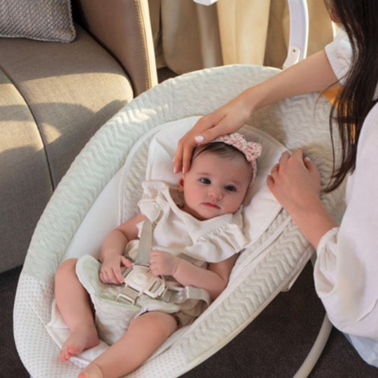 Baby in a white bassinet with a woman adjusting the straps, sitting on a couch.