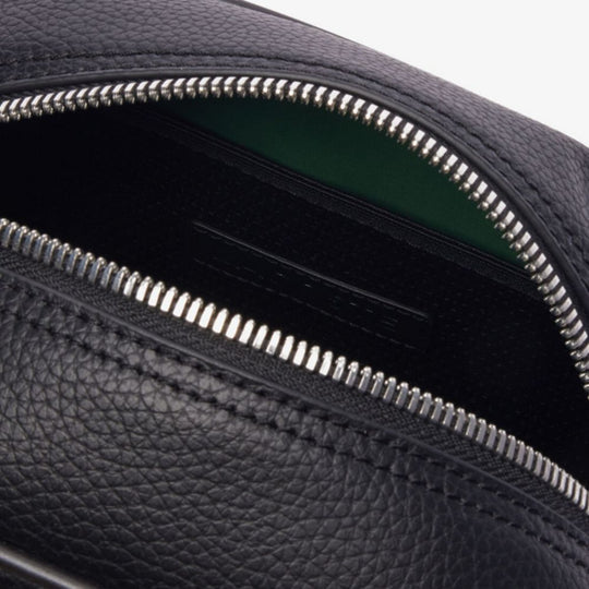 Close-up shot of the open LACOSTE Angy Mesh Pocket Vanity Bag, revealing its black interior with a glimpse of a bright green lining deeper inside. The bag's exterior is made of black, textured grain leather, and a silver-toned zipper with visible teeth runs along the opening.