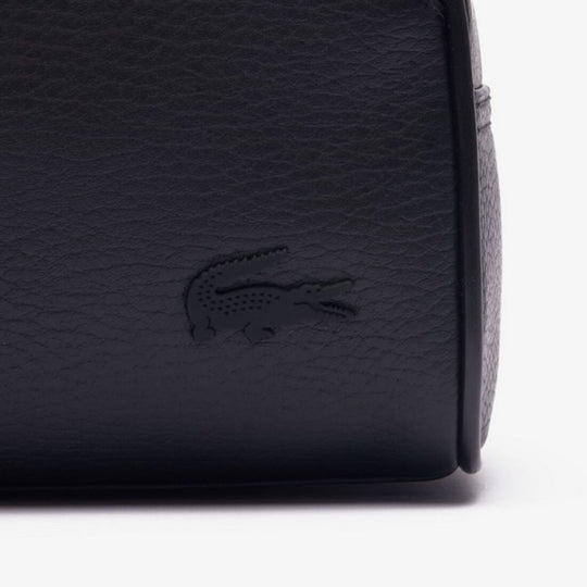 Close-up shot of the LACOSTE Angy Mesh Pocket Vanity Bag, prominently featuring the tonal, debossed crocodile logo on its black, pebbled grain leather surface. The image shows the side and bottom curvature of the bag.
