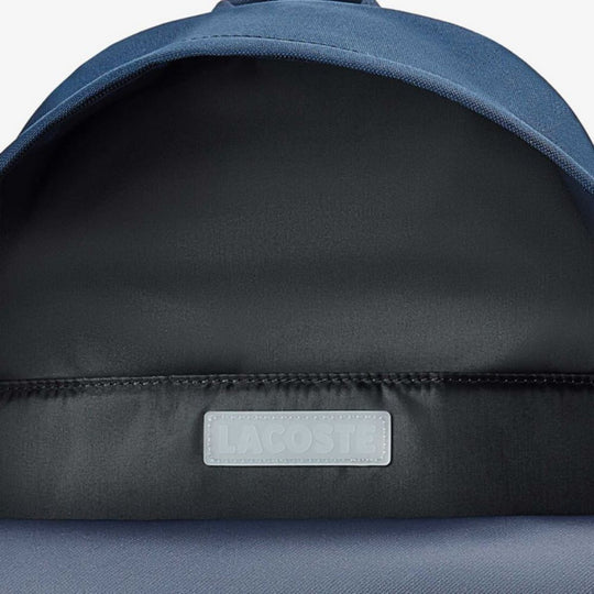 A close-up shot of the interior of a dark blue Lacoste backpack. The main compartment is open, revealing a black lining with a white rectangular Lacoste logo patch sewn onto it. The top curved portion of the backpack is visible, showing the smooth, dark blue textile material.