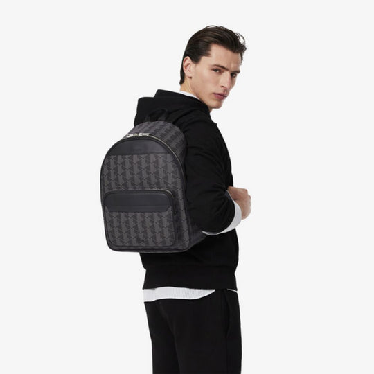 Man from behind wearing a black hoodie and sweatpants, carrying a Lacoste The Blend Backpack with a dark monogram pattern and black accents, looking back at the viewer.