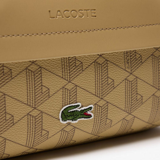 A close-up of the Lacoste The Blend Grained Texture Vanity Bag, showcasing its beige monogram pattern and a prominent embroidered green crocodile logo on the front. Above the pattern, a smooth beige leather-like strip features the embossed "LACOSTE" text.