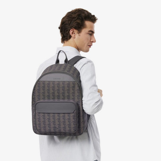 A person, viewed from the back and slightly to the right, wears a light-colored striped shirt and carries a Lacoste The Blend Backpack on their back. The backpack has a grey and black geometric monogram pattern with solid grey accents and features a front zip pocket. The person is looking over their right shoulder.