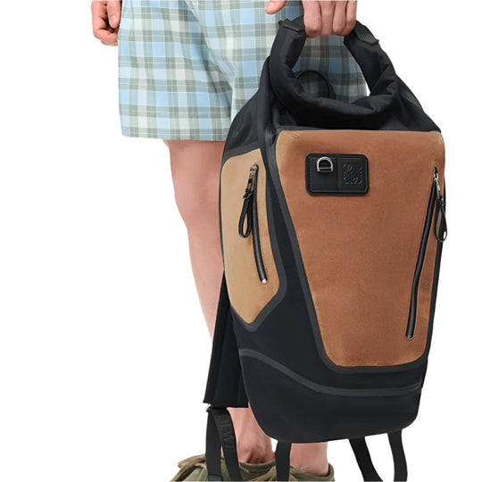 A person holds a LOEWE Flex Roll-Top Backpack by its top handle, against a white background.  He is wearing light blue and white checkered shorts and part of a green shoe is visible at the bottom.