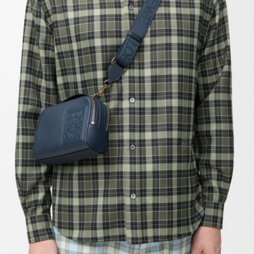 Person wearing a green plaid shirt with  LOEWE Mini Crossbody Camera Bag in Satin Calfskin over one shoulder on a white background