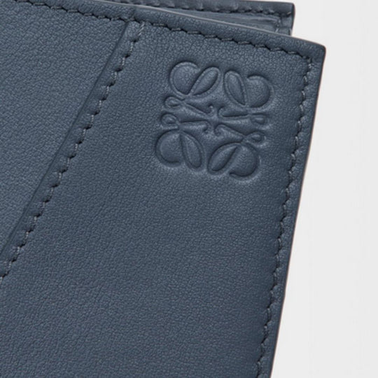A close-up shot of a blue leather bifold wallet, featuring the embossed LOEWE Anagram logo on the bottom right corner of the visible panel. The texture of the classic calfskin leather and the neat stitching along the edges are clearly visible.