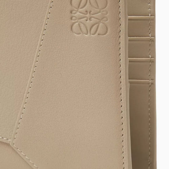 A close-up of a beige LOEWE Puzzle Edge Bi-fold Wallet in classic calfskin, showing the textured, geometric "puzzle" stitching and the embossed Anagram logo.