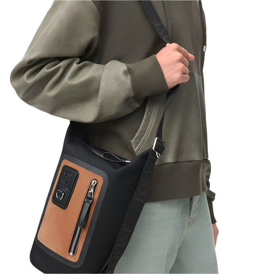 A person, from the waist up, wears a dark olive green hoodie with lighter green patches on the shoulders and arms, paired with light wash jeans. They are holding the black LOEWE Small Flex Loop Bucket bag by its black LOEWE-branded strap, which is slung over their shoulder.