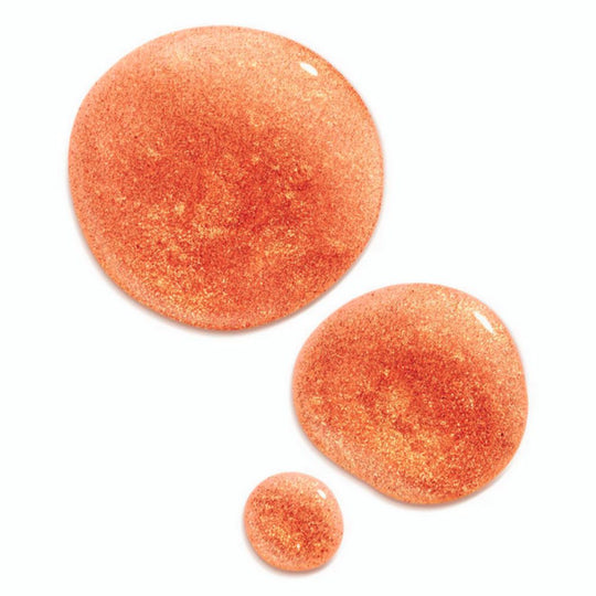 Three glistening drops of shimmering body oil are arranged on a white background, varying in size from largest to smallest. The oil is a warm, coppery orange color, infused with fine gold glitter particles that catch the light. The largest drop is at the top left, a medium-sized drop is below and to its right, and the smallest drop is at the bottom center.