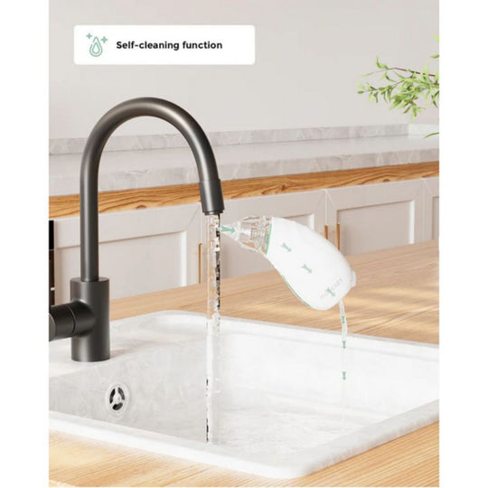 A Momcozy Quiet Rechargeable Baby Nasal Aspirator is being cleaned under a running faucet in a kitchen sink, demonstrating its self-cleaning function. The device's tip is positioned under the stream of water, which flows through the aspirator and out into the sink.