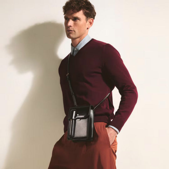 Man wearing a burgundy sweater and brown pants with a black crossbody bag against a beige background