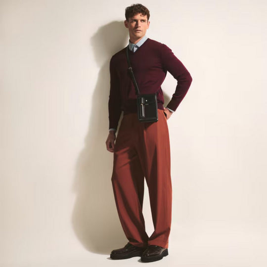 A man wearing a burgundy V-neck sweater over a light blue collared shirt and rust-colored wide-leg pants stands against a white wall, looking to his right. A black Montblanc small messenger bag with a pen case attached is slung across his body.