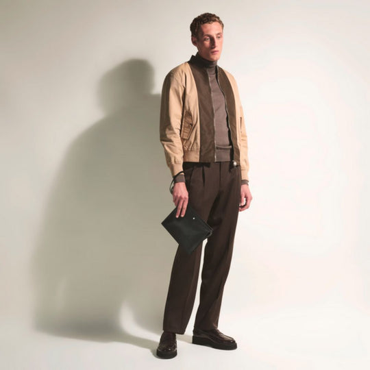 Man wearing a beige jacket and brown pants holding a black clutch against a plain background