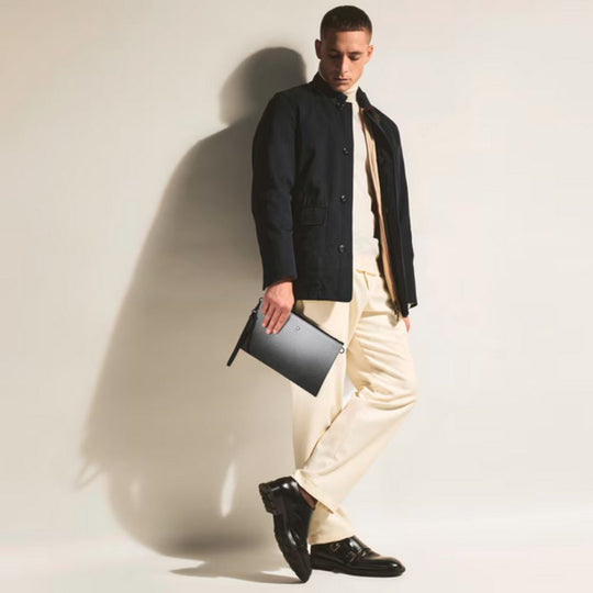 Man wearing a black coat, white shirt, and beige pants holding Montblanc Extreme 3.0 Lightweight Stylish Cross-Body Pouch against a plain background