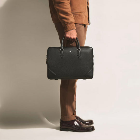 A person in a brown suit and burgundy loafers stands, holding a black Montblanc grain leather document case with a silver emblem.