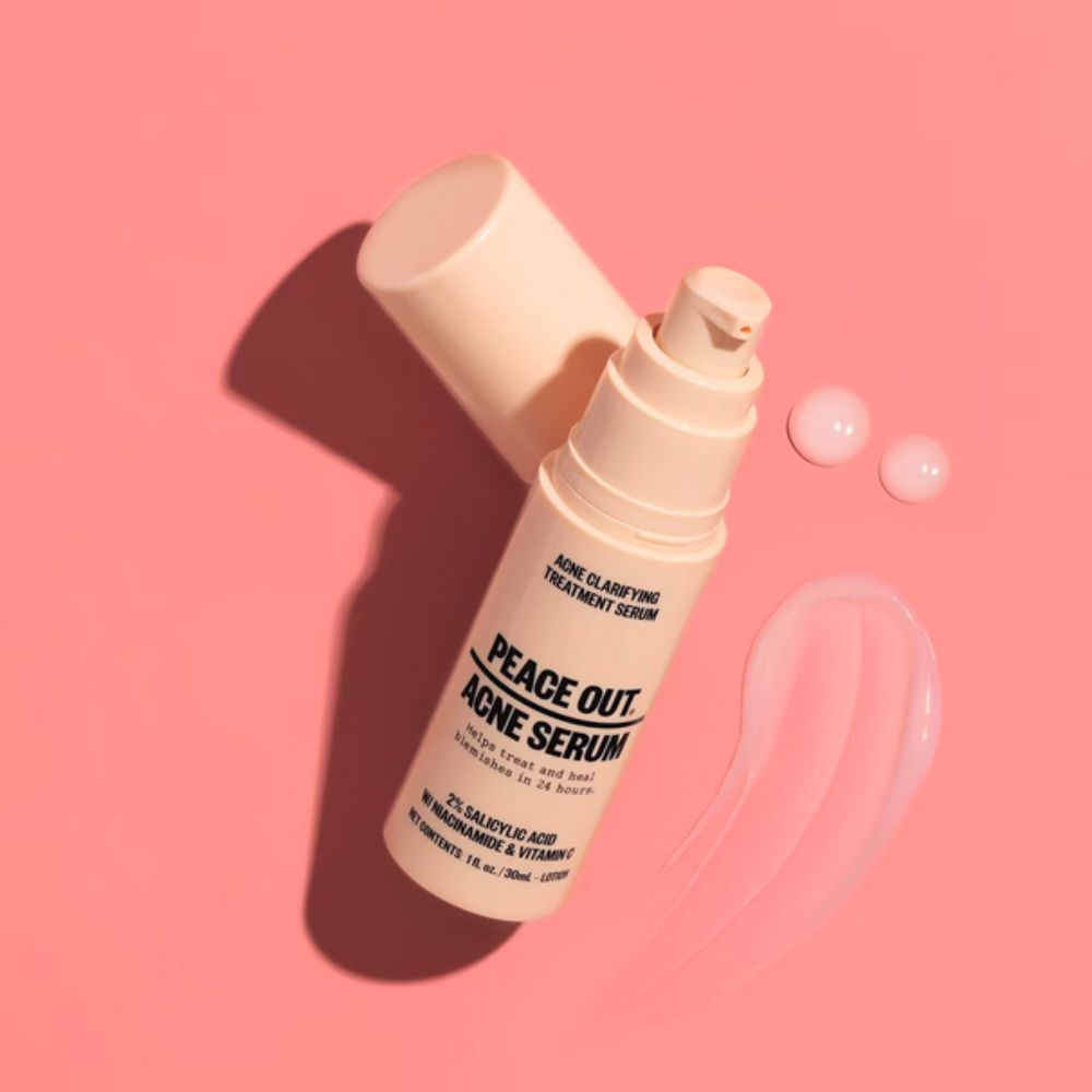 A light peach-colored pump bottle of Peace Out Acne Serum sits on a pink background, with its cap removed and two drops of serum alongside a smear of product.