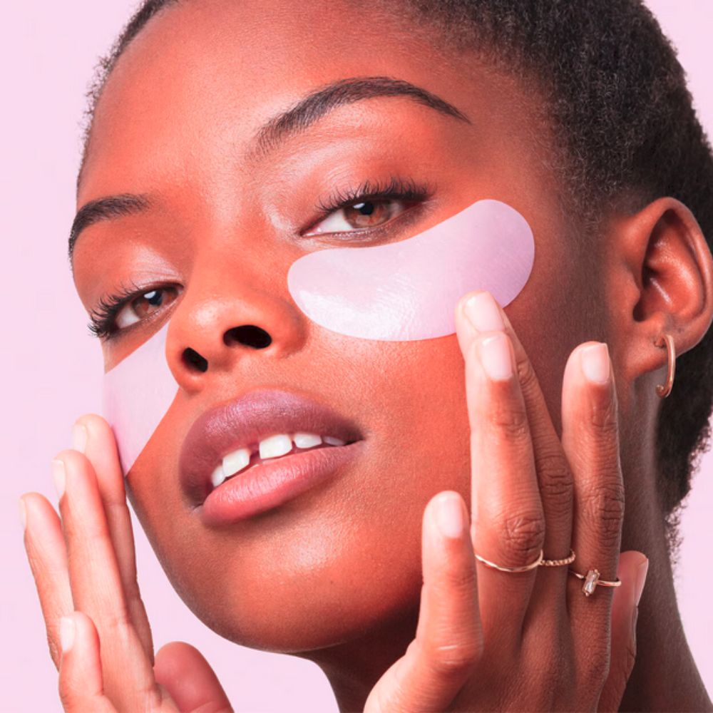 A person with brown skin and dark hair looks up and to the right, touching pink under-eye patches with both hands. These eyes patches are Peace Out Skincare Puffy Eyes - Rejuvinating Under-Eye Patches