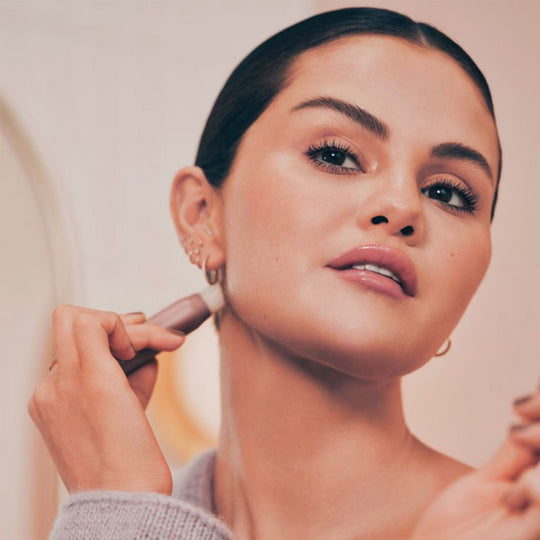 Selena Gomez, with hair pulled back, applies a light-toned Rare Beauty Find Comfort Stop & Soothe Aromatherapy Pen to her neck near her ear, looking up with a soft expression in a warm, softly lit setting.