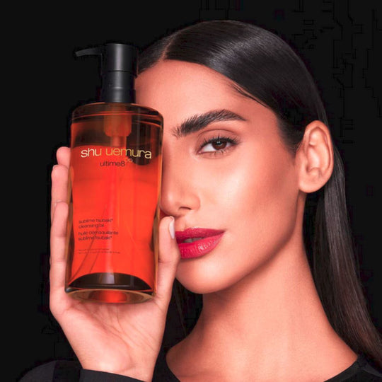 A woman partially obscures her face with a bottle of Shu Uemura ultime8 sublime beauty cleansing oil