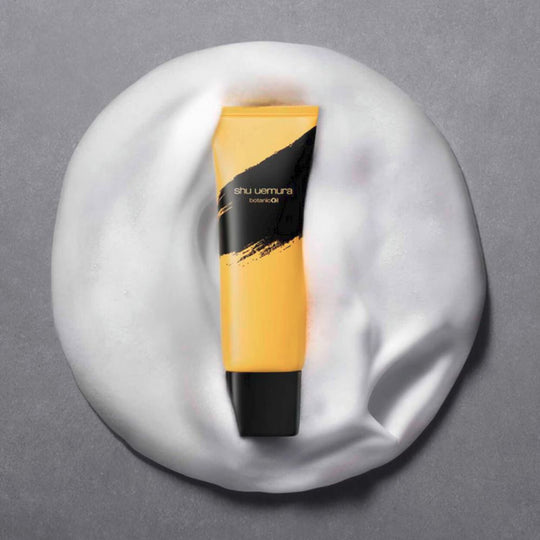 A yellow and black tube of Shu Uemura Botanicoil Gentle Amino Cleansing Foam is displayed on a bed of white foam against a gray background.
