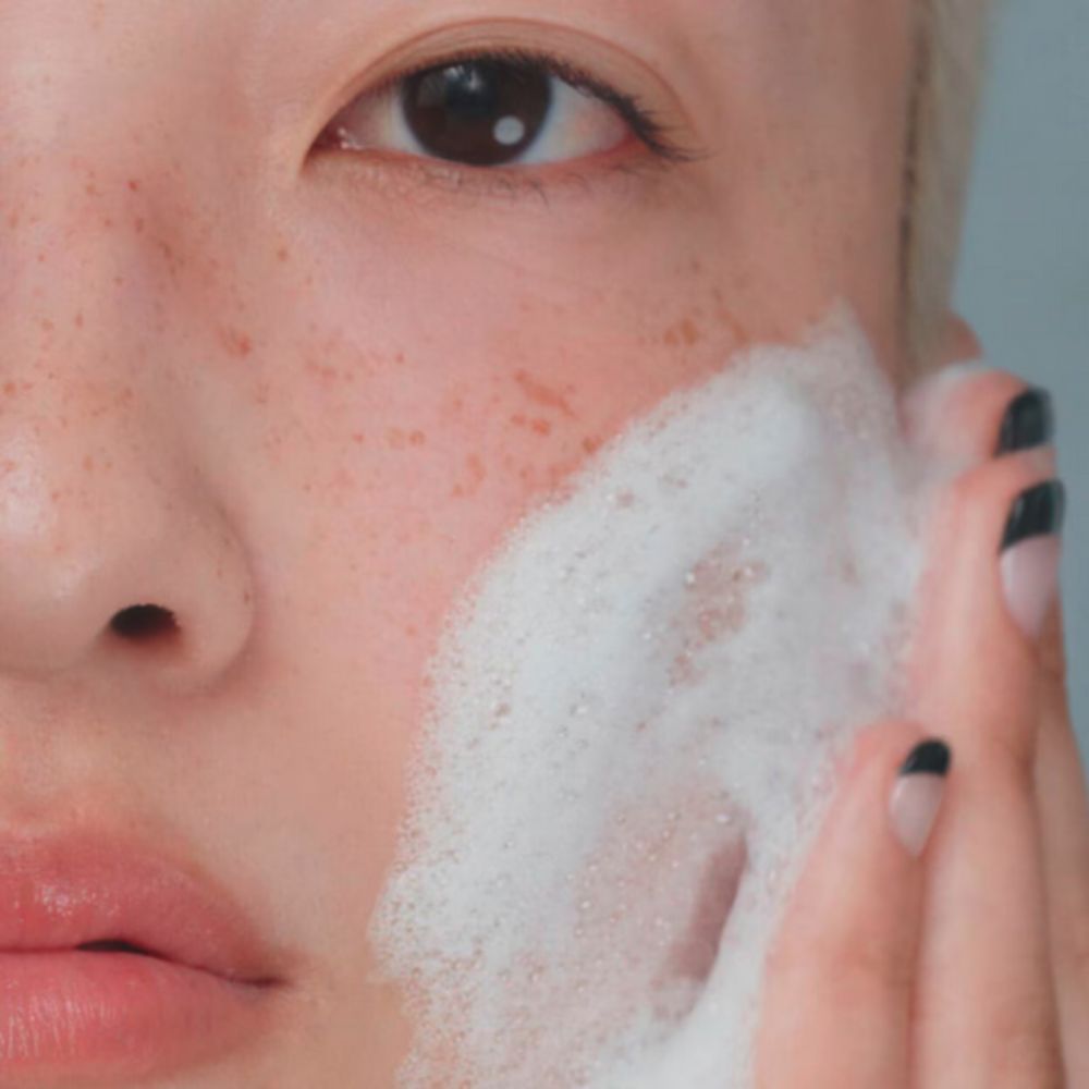 A close-up of a person's face with light skin and freckles, applying white foam to their cheek. One eye is visible, looking towards the viewer, and fingers with black and nude nail polish are near the cheek
