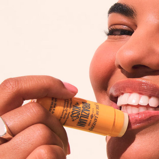 Close-up of a person applying an orange SOL DE JANEIRO Brazilian Kiss lip product, with visible text on the tube and a dewy shine on the lips.