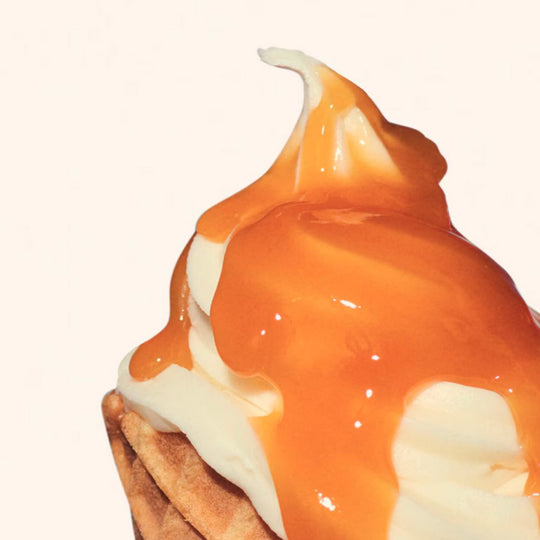 A swirl of soft-serve ice cream in a waffle cone is drizzled with a thick, glossy caramel or dulce de leche sauce.