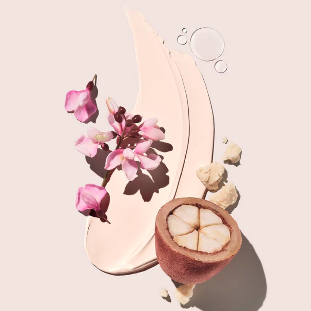 A close-up composition on a pale background featuring a swatch of light-colored lotion, pink flowers on a stem, a halved fruit (likely cupuaçu), irregular chunks of a solid substance (like butter), and clear liquid droplets.