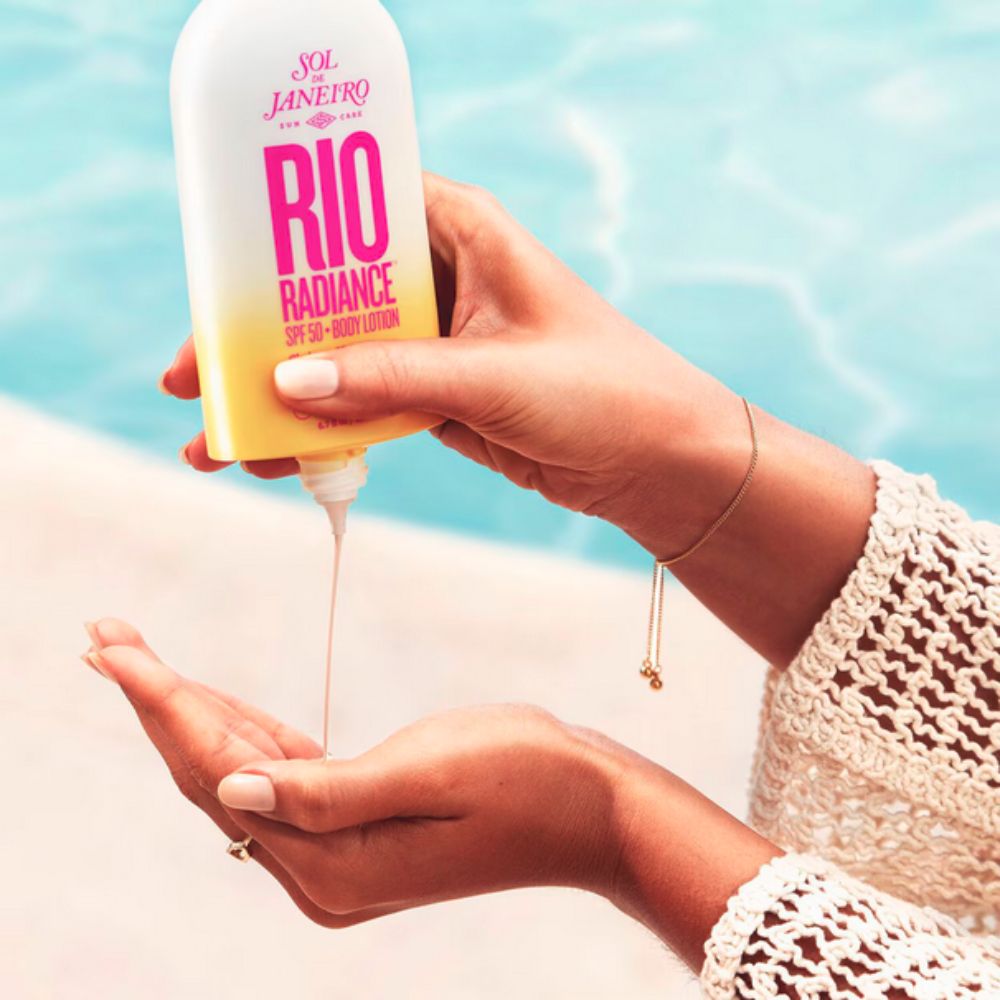 Hands are shown pouring the Sol de Janeiro Rio Radiance SPF 50 Body Lotion from its bottle into a palm, with a blurred blue water background.