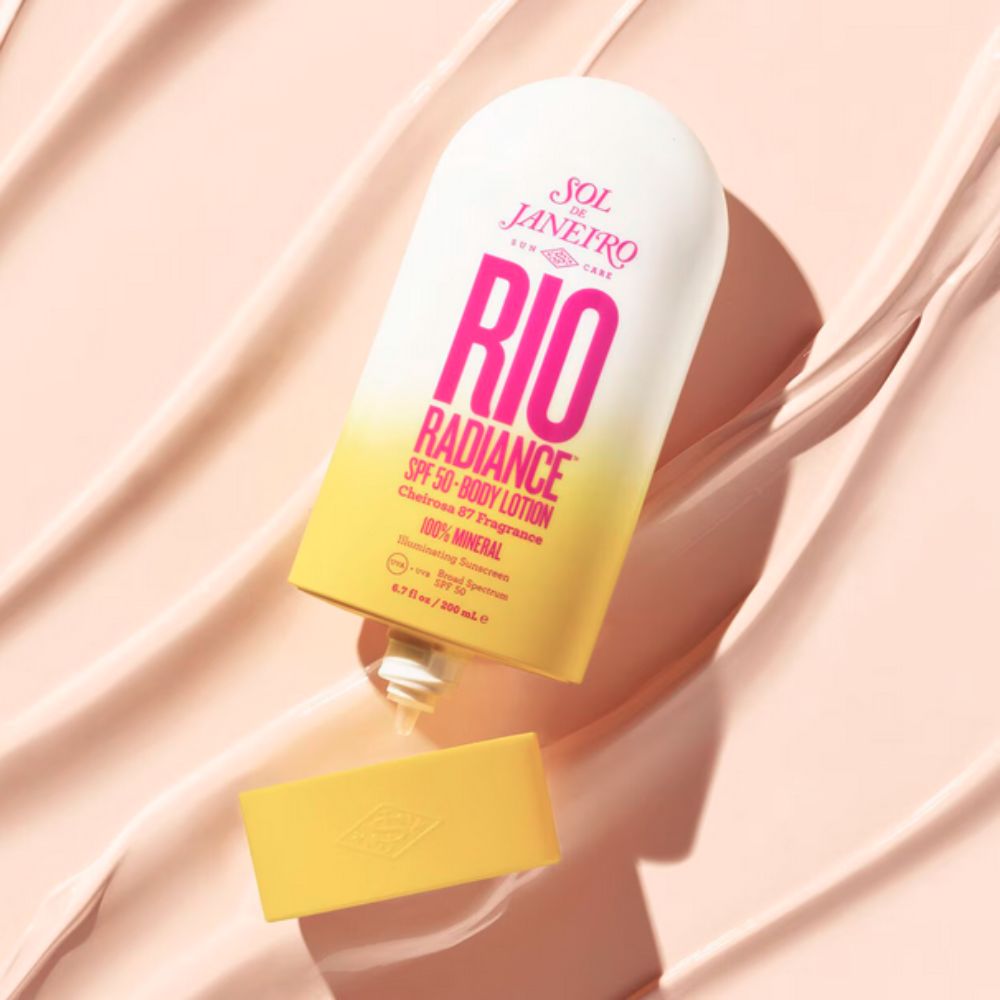 The bottle of Sol de Janeiro Rio Radiance SPF 50 Body Lotion is shown lying on its side on a textured, creamy background