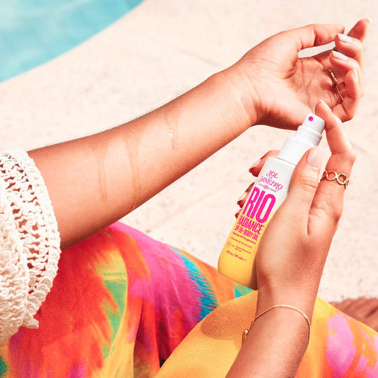 Person spraying SOL DE JANEIRO Rio Radiance SPF 50 Body Oil onto their arm next to a pool, showing the oil glistening on the skin.