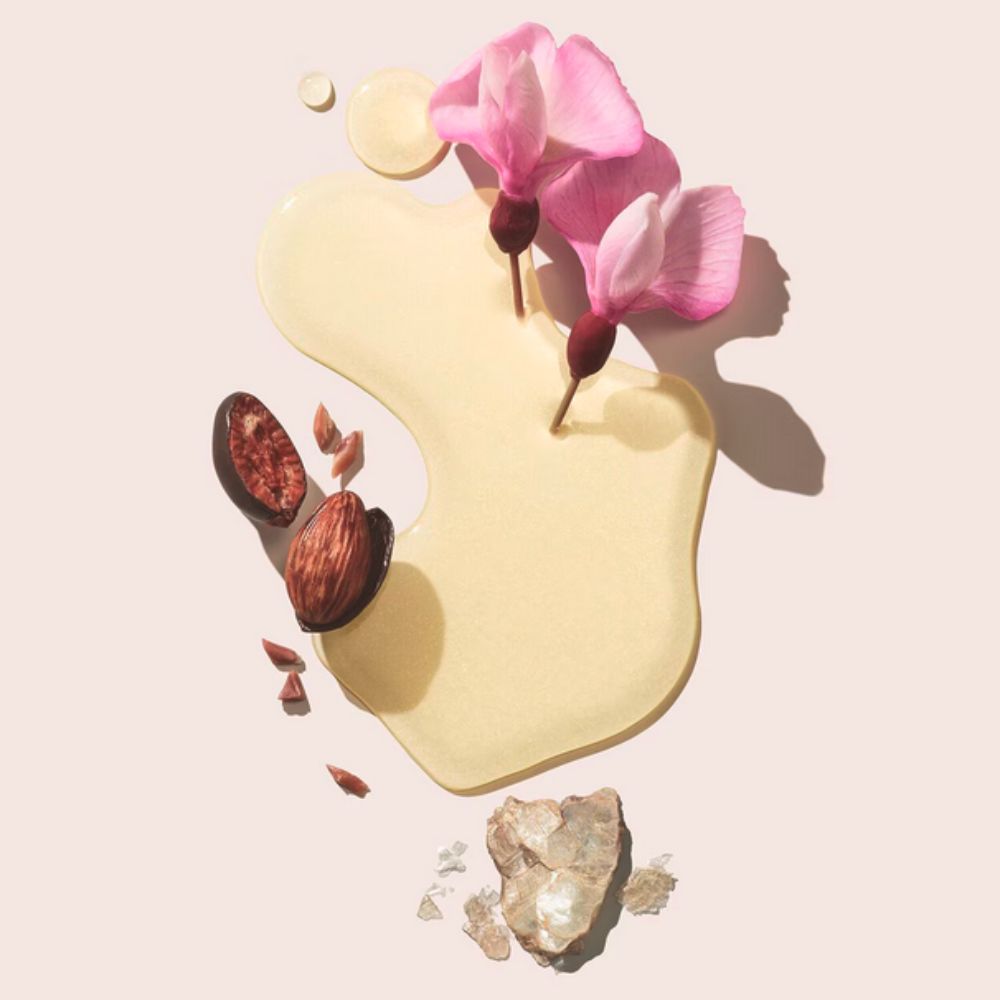 Flat lay of a pool of yellow oil, pink flowers, seeds/pods, and a crystal, representing body oil ingredients.